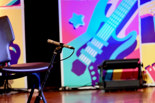 A dynamic, colorful background featuring a stylized guitar in blues and purples with abstract shapes in the background. In the foreground, there's an empty chair and a microphone on a stand, indicating a performance setting.