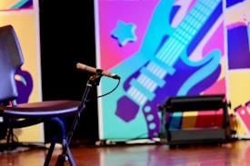 A dynamic, colorful background featuring a stylized guitar in blues and purples with abstract shapes in the background. In the foreground, there's an empty chair and a microphone on a stand, indicating a performance setting.