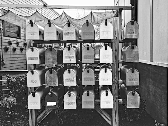 A collection of metal mailboxes is neatly arranged in several rows and columns on a wooden stand. Each mailbox is labeled with a number, indicating addresses or apartment units. The setting is outdoors, with some plants visible along the side of a building.