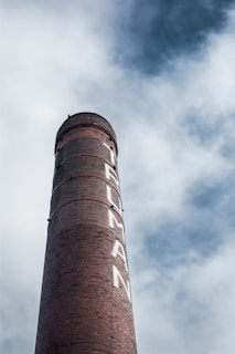 A tall brick chimney with the word 'HUMAN' painted vertically on its side. The chimney rises against a backdrop of cloudy sky, creating a striking contrast between the industrial structure and nature.