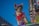 A small dog wearing colorful attire, including a red cap and goggles, stands against a vivid blue sky. It is adorned with a red bow and multicolored decorations, giving a festive appearance. The background includes blurred urban elements and bright, colorful lights, suggesting a parade or celebration setting.