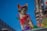 A small dog wearing colorful attire, including a red cap and goggles, stands against a vivid blue sky. It is adorned with a red bow and multicolored decorations, giving a festive appearance. The background includes blurred urban elements and bright, colorful lights, suggesting a parade or celebration setting.