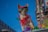 A small dog wearing colorful attire, including a red cap and goggles, stands against a vivid blue sky. It is adorned with a red bow and multicolored decorations, giving a festive appearance. The background includes blurred urban elements and bright, colorful lights, suggesting a parade or celebration setting.