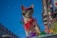 A small dog wearing colorful attire, including a red cap and goggles, stands against a vivid blue sky. It is adorned with a red bow and multicolored decorations, giving a festive appearance. The background includes blurred urban elements and bright, colorful lights, suggesting a parade or celebration setting.