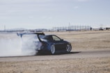 A dark-colored sports car is drifting, creating a large cloud of white smoke behind it. The car has a large rear spoiler and is on a flat, dry terrain, possibly a racetrack. There are mountains and a clear blue sky in the background.