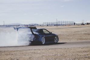 A dark-colored sports car is drifting, creating a large cloud of white smoke behind it. The car has a large rear spoiler and is on a flat, dry terrain, possibly a racetrack. There are mountains and a clear blue sky in the background.