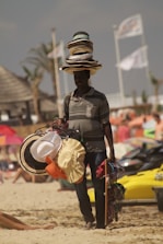 A person selling hats on a beach with several hats stacked on their head, while holding additional hats and accessories. The background shows a beach setting with some blurred people and beach elements like umbrellas and jet skis.