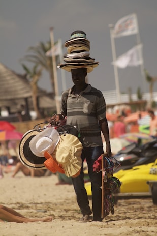 A person selling hats on a beach with several hats stacked on their head, while holding additional hats and accessories. The background shows a beach setting with some blurred people and beach elements like umbrellas and jet skis.