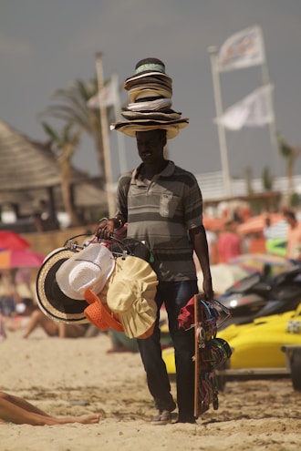 A person selling hats on a beach with several hats stacked on their head, while holding additional hats and accessories. The background shows a beach setting with some blurred people and beach elements like umbrellas and jet skis.