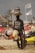 A person selling hats on a beach with several hats stacked on their head, while holding additional hats and accessories. The background shows a beach setting with some blurred people and beach elements like umbrellas and jet skis.