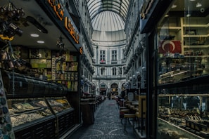 A narrow, covered shopping arcade with a high arched glass roof. The space is flanked by historic buildings and various shops on both sides, displaying colorful goods in the windows. On the left, there are shelves filled with snacks and delicacies, while on the right, there are souvenirs and a Turkish flag displayed. The ground is paved with cobblestones, adding to the historic ambiance. Soft lighting creates a cozy and inviting atmosphere.