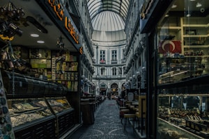 A narrow, covered shopping arcade with a high arched glass roof. The space is flanked by historic buildings and various shops on both sides, displaying colorful goods in the windows. On the left, there are shelves filled with snacks and delicacies, while on the right, there are souvenirs and a Turkish flag displayed. The ground is paved with cobblestones, adding to the historic ambiance. Soft lighting creates a cozy and inviting atmosphere.