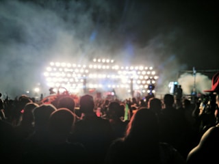 A large crowd of people is gathered outdoors in front of a brightly lit stage, creating an atmosphere of excitement and anticipation. The stage is filled with numerous lights, casting a glow and creating dramatic contrasts with the night sky. The image captures the silhouettes of the audience, emphasizing the event's energy and scale.