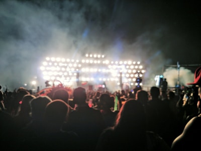 A large crowd of people is gathered outdoors in front of a brightly lit stage, creating an atmosphere of excitement and anticipation. The stage is filled with numerous lights, casting a glow and creating dramatic contrasts with the night sky. The image captures the silhouettes of the audience, emphasizing the event's energy and scale.