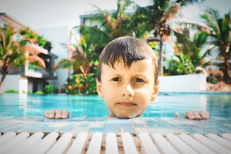 A young child with wet hair is holding on to the edge of a swimming pool. In the background, there are palm trees and buildings, suggesting a tropical or resort setting. The water in the pool is a bright blue, and the sunlight creates a warm and inviting atmosphere.