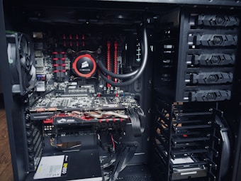 An open computer case with visible internal components, including a cooling fan, a Corsair liquid cooler, MSI graphics cards, RAM modules, and a power supply unit. The components are neatly arranged with power and data cables connected to different parts.