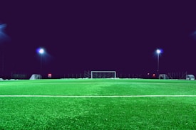 An empty artificial turf soccer field is illuminated by bright stadium lights at night. A goalpost is located in the center background with additional goalposts and various equipment placed around the field.