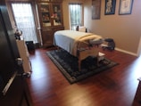 A well-organized room features a massage table covered with a white sheet and a folded towel at one end, situated on a decorative rug with intricate patterns. The wooden floor adds warmth to the ambiance. Against the far wall stands an ornately carved wooden cabinet displaying various items. There are two tall windows covered with bamboo shades allowing soft natural light to filter through. Several framed artworks hang on the adjacent wall. A small black stool is placed near the massage table.