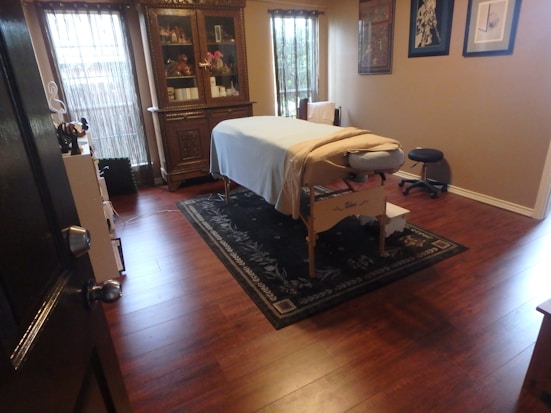 A well-organized room features a massage table covered with a white sheet and a folded towel at one end, situated on a decorative rug with intricate patterns. The wooden floor adds warmth to the ambiance. Against the far wall stands an ornately carved wooden cabinet displaying various items. There are two tall windows covered with bamboo shades allowing soft natural light to filter through. Several framed artworks hang on the adjacent wall. A small black stool is placed near the massage table.