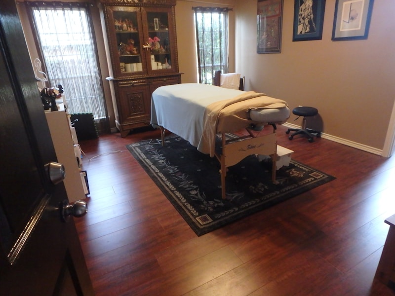 A well-organized room features a massage table covered with a white sheet and a folded towel at one end, situated on a decorative rug with intricate patterns. The wooden floor adds warmth to the ambiance. Against the far wall stands an ornately carved wooden cabinet displaying various items. There are two tall windows covered with bamboo shades allowing soft natural light to filter through. Several framed artworks hang on the adjacent wall. A small black stool is placed near the massage table.