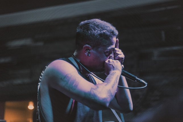 A person is performing on stage with intense focus, holding a microphone close to their mouth. The setting appears dimly lit, and the performer exhibits tattoos on their arm and short hair, exuding a sense of passion and engagement.
