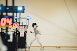 A fencer in full fencing gear, including a helmet and white protective clothing, is poised and lunging forward on a piste. In the background, electronic scoring equipment displays numbers and time. The scene suggests an ongoing fencing match in a sports hall.