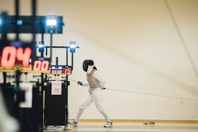 A fencer in full fencing gear, including a helmet and white protective clothing, is poised and lunging forward on a piste. In the background, electronic scoring equipment displays numbers and time. The scene suggests an ongoing fencing match in a sports hall.