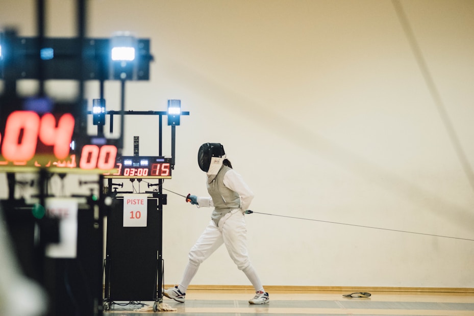 A fencer in full fencing gear, including a helmet and white protective clothing, is poised and lunging forward on a piste. In the background, electronic scoring equipment displays numbers and time. The scene suggests an ongoing fencing match in a sports hall.