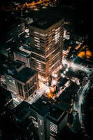 An aerial view of a city at night, with several tall buildings illuminated by artificial lights. Streets are visible along with light trails from moving vehicles, creating a vibrant urban scene.
