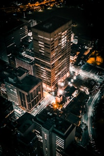 An aerial view of a city at night, with several tall buildings illuminated by artificial lights. Streets are visible along with light trails from moving vehicles, creating a vibrant urban scene.