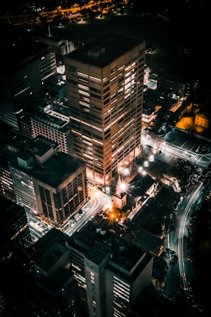 An aerial view of a city at night, with several tall buildings illuminated by artificial lights. Streets are visible along with light trails from moving vehicles, creating a vibrant urban scene.