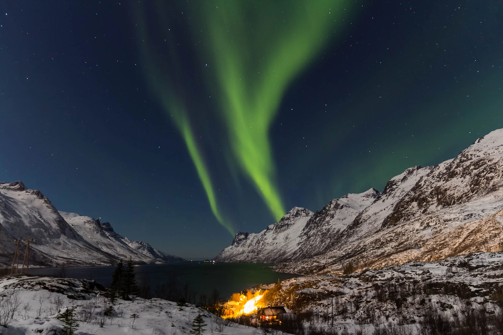 Green aurora curtains above a snow-filled fjord and a single lit cabin at Ersfjord near Tromsø