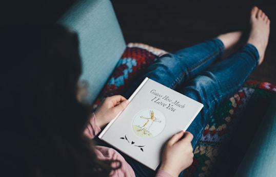 A person is sitting comfortably on a couch, holding a children's book titled 'Guess How Much I Love You.' The book cover features an illustration of two rabbits embracing. The person is dressed in casual clothing, including a pink top and blue jeans, and their feet are bare. A multicolored knit blanket partially covers the couch.