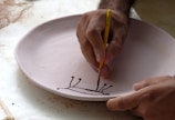 A person is delicately painting a design on a circular, unglazed ceramic plate with a fine brush. The design appears to be abstract or floral, created with a dark brown paint. The hands are focused and steady, indicating a careful and precise painting process.