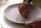 A person is delicately painting a design on a circular, unglazed ceramic plate with a fine brush. The design appears to be abstract or floral, created with a dark brown paint. The hands are focused and steady, indicating a careful and precise painting process.