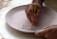 A person is delicately painting a design on a circular, unglazed ceramic plate with a fine brush. The design appears to be abstract or floral, created with a dark brown paint. The hands are focused and steady, indicating a careful and precise painting process.