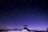 Under a vast, starry sky, an observatory stands silhouetted against a twilight backdrop. The scene captures a serene, night-time moment with a single person standing by the structure. The rich gradient of blue to purple across the sky adds depth and tranquility.