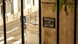 A narrow, sunlit street with a pedestrian crossing and road signs. A wall with a sign reading 'Travessa de São Tomé' is in the foreground, partially obscured by iron bars. Green foliage hangs in from the top right corner.