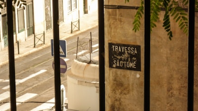 A narrow, sunlit street with a pedestrian crossing and road signs. A wall with a sign reading 'Travessa de São Tomé' is in the foreground, partially obscured by iron bars. Green foliage hangs in from the top right corner.