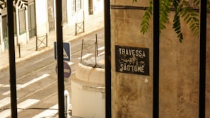 A narrow, sunlit street with a pedestrian crossing and road signs. A wall with a sign reading 'Travessa de São Tomé' is in the foreground, partially obscured by iron bars. Green foliage hangs in from the top right corner.