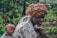 A woman is carrying a sleeping baby on her back using a patterned cloth wrap. She wears a vibrant, multicolored head wrap and a floral-patterned blouse. The background is a lush, green environment that resembles a natural or rural setting.