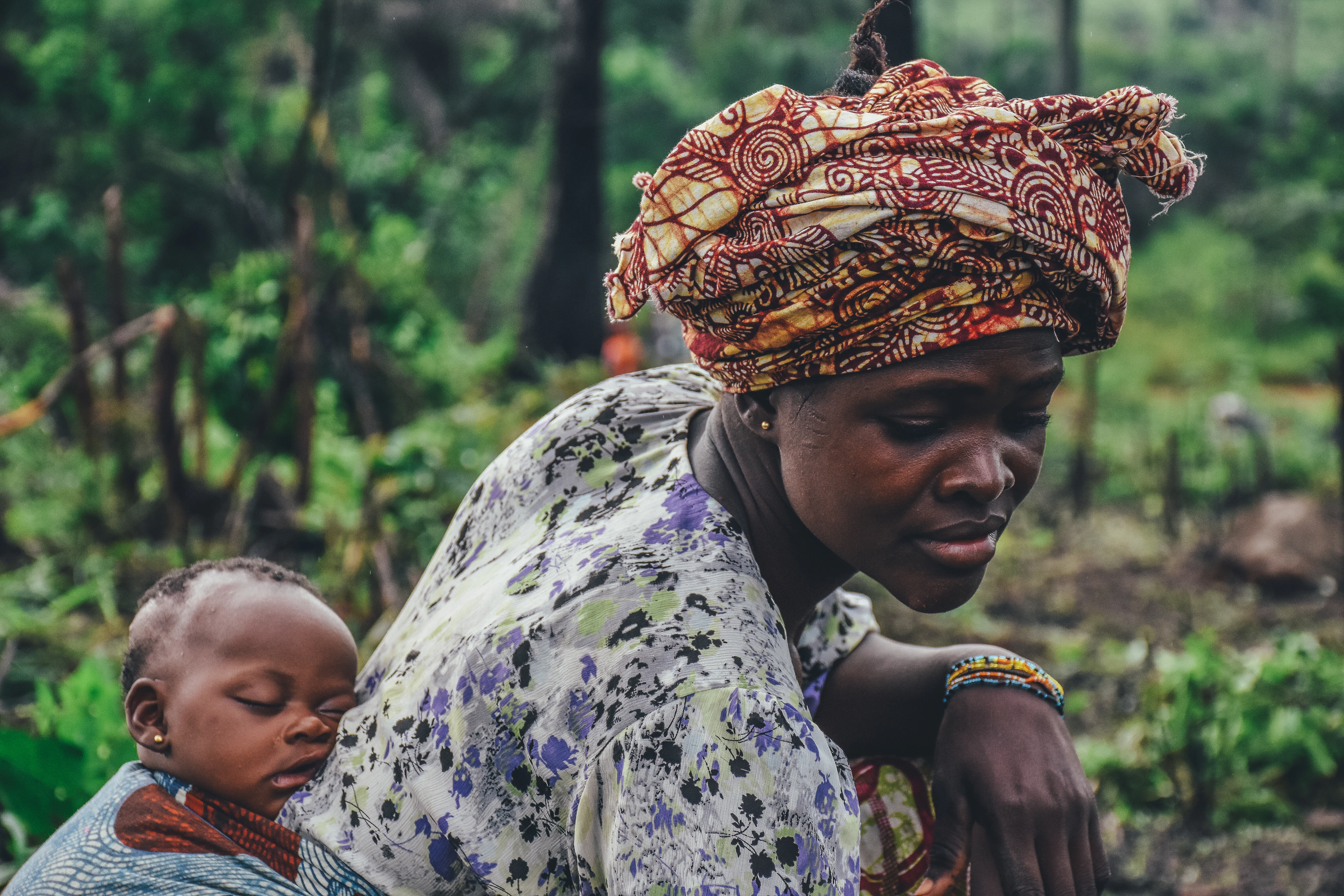 A woman in a patterned head wrap carrying a child on her back, surrounded by greenery