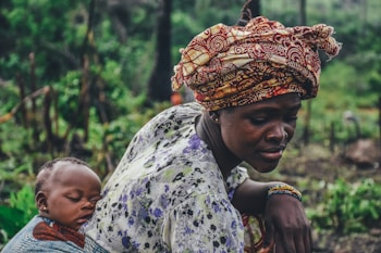 A woman is carrying a sleeping baby on her back using a patterned cloth wrap. She wears a vibrant, multicolored head wrap and a floral-patterned blouse. The background is a lush, green environment that resembles a natural or rural setting.