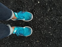 A pair of bright turquoise running shoes worn by someone in jeans, standing on a rough, textured surface of small, dark stones or gravel.
