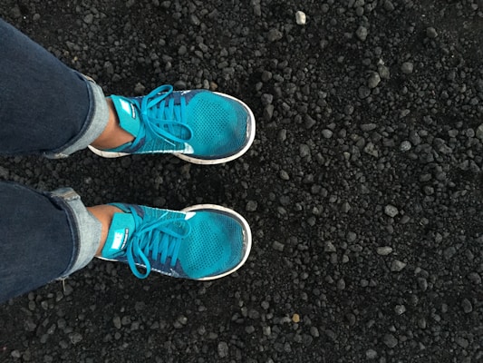A pair of bright turquoise running shoes worn by someone in jeans, standing on a rough, textured surface of small, dark stones or gravel.