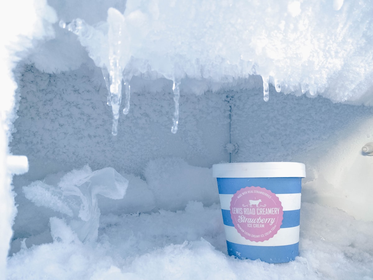 Interior de un freezer cubierto de escarcha gruesa y estalactitas de hielo colgando del techo, con un pote de helado de frutilla adentro