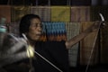 An elderly person is engaged in traditional weaving. The background features a variety of colorful, intricately patterned textiles. The person's expression appears concentrated, and their hands are actively involved in the weaving process.
