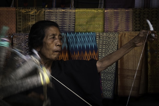 An elderly person is engaged in traditional weaving. The background features a variety of colorful, intricately patterned textiles. The person's expression appears concentrated, and their hands are actively involved in the weaving process.
