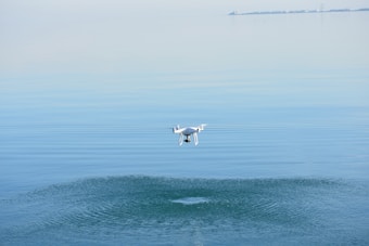 A white drone hovers above a serene body of water, creating gentle ripples on the surface. The background showcases a calm horizon with a slight gradient in the sky, and distant land can be faintly seen.