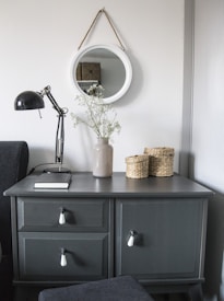 A gray cabinet with three drawers is set against a white wall. Above it hangs a round mirror with a cord. The cabinet top holds a beige vase with delicate white flowers, a black desk lamp, a closed book, and two small woven baskets.